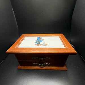 Puede incluir: Una caja de joyería de madera con dos cajones y una parte superior decorativa. La parte superior presenta un azulejo blanco con una ilustración colorida de un niño y un muñeco de nieve. La caja tiene tiradores de cajón de metal ornamentados y un acabado marrón cálido.