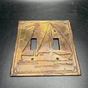 May include: A square brass-toned metal double light switch wall plate featuring an embossed design of a sailboat on water with a bird in the sky. The plate has two rectangular cutouts for switches and four small circular screw holes.