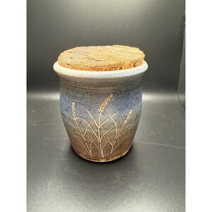 May include: A ceramic jar with a cork lid. The jar is blue and brown with etched wheat stalks. The rim is a lighter blue. The cork lid is a light brown colour. The jar is a decorative storage container.