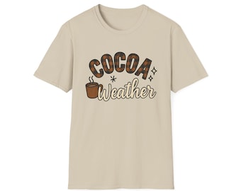May include: A beige t-shirt featuring the words "COCOA Weather" in a brown, retro font. The word "COCOA" has a plaid pattern, and a steaming mug of cocoa is depicted below the text. The shirt has short sleeves and a crew neck.