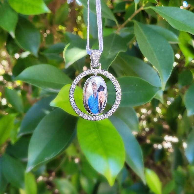 May include: A silver pendant necklace with a photo of two people inside an oval frame. The frame is surrounded by a circle of small, clear crystals. The necklace hangs from a thin, white ribbon against a backdrop of green leaves.