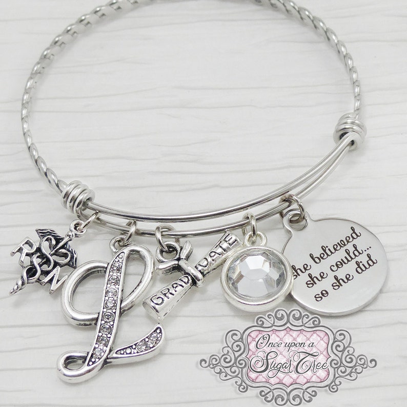 May include: Silver-toned charm bracelet with multiple dangles. Charms include an "L" encrusted with rhinestones, a graduation scroll, a clear crystal, and a round charm with the inscription "she believed she could... so she did."