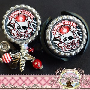 May include: Two badge reels with a pirate nurse skull design. The skull wears a red nurse's cap and an eye patch. The text "ARRRR TEEEE!!!" and "AMBER" are visible. One reel has a charm with beads and a medical symbol.