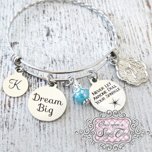 May include: A silver charm bracelet with a variety of charms, including a charm that says "Dream Big", a charm that says "Never Let Anyone Dull Your Sparkle", a charm with the letter "K", a charm with the number "21", and a blue crystal charm. The bracelet is on a white wooden background.
