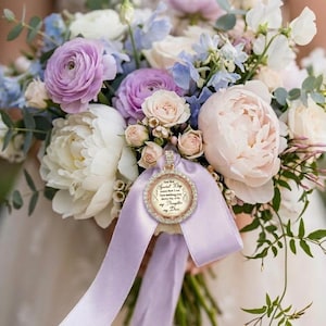 May include: A wedding bouquet of white and lavender peonies, roses, and blue delphiniums features a circular photo charm with a rhinestone border and a light purple ribbon. The charm displays text that reads: On my Special Day, love that I am here making one dream the only my Daughter my Day.