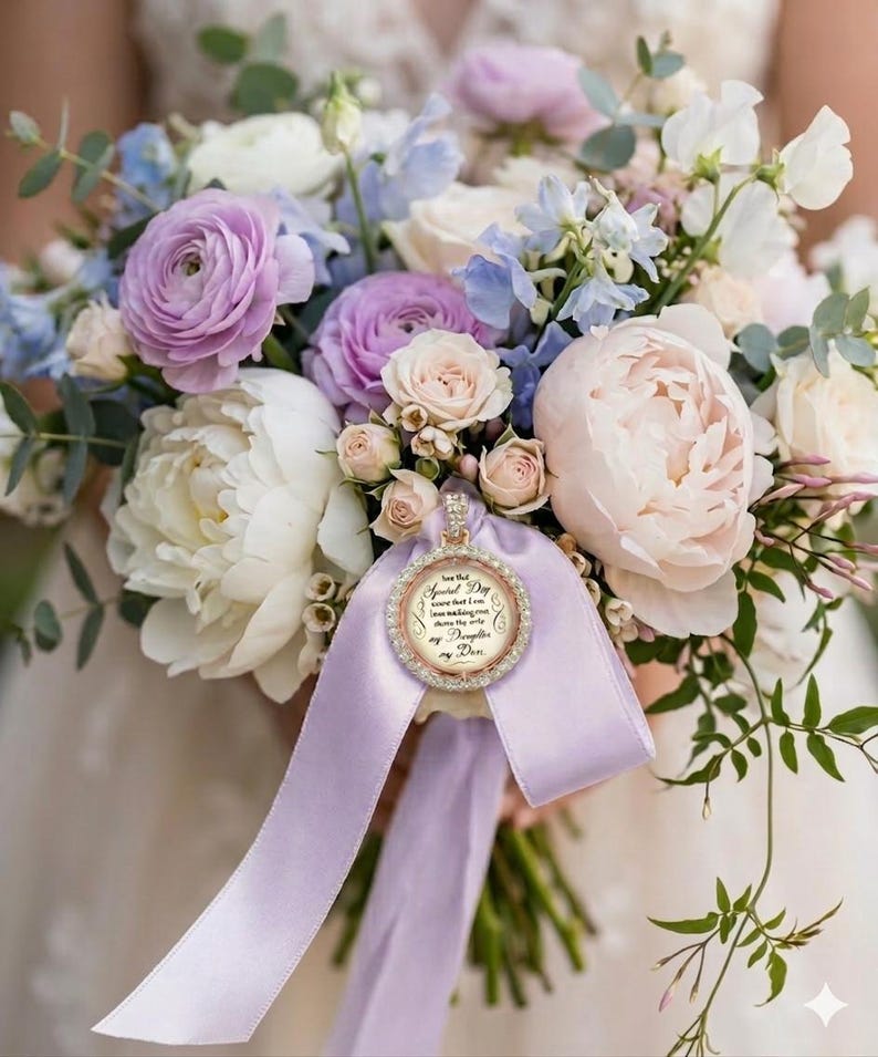 May include: A wedding bouquet of white and lavender peonies, roses, and blue delphiniums features a circular photo charm with a rhinestone border and a light purple ribbon. The charm displays text that reads: On my Special Day, love that I am here making one dream the only my Daughter my Day.