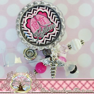 May include: A pink and black patterned badge reel with a silver cap featuring a pink heart with the letters RT and the name Kathleen. The reel has a silver retractable cord and a silver charm with a medical caduceus symbol and wings.