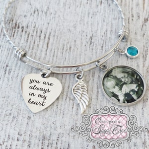 May include: A silver bangle bracelet with a heart charm that says "you are always in my heart", a silver angel wing charm, a round photo charm, and a turquoise blue crystal charm. The bracelet is on a wooden surface.