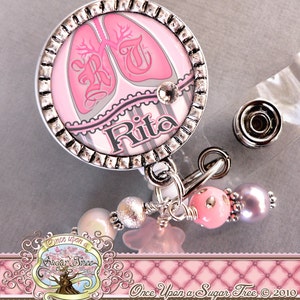 May include: A pink and silver badge holder with a picture of lungs and the name "Rita" on it. The badge holder has a silver chain with pink and silver beads attached to it.