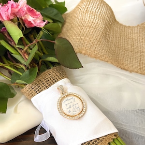 May include: A round gold-toned pendant featuring a border of clear crystals and a central white insert with the black script text "Missing you as I walk down the aisle," attached to a white fabric-wrapped bouquet with burlap accents.