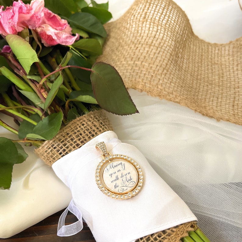 May include: A round gold-toned pendant featuring a border of clear crystals and a central white insert with the black script text "Missing you as I walk down the aisle," attached to a white fabric-wrapped bouquet with burlap accents.