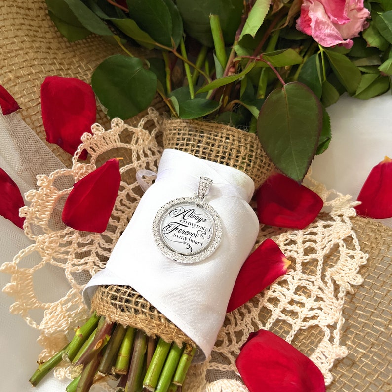 May include: A white bouquet of red roses with a silver charm attached. The charm has a clear crystal border and the words "Always on my mind Forever in my heart" written on it.