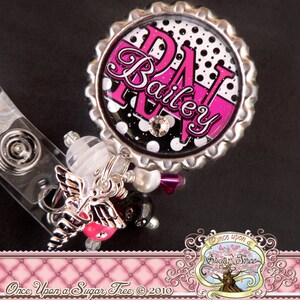 May include: A pink and black polka dot badge reel with the initials "D.A." and the name "Bailey" in a pink script font. The reel is attached to a silver charm with a white pearl and a silver angel wing.