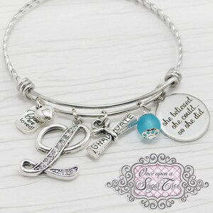 May include: Silver-toned charm bracelet with multiple pendants. Charms include a rhinestone-studded letter "L", a graduation scroll, a blue bead, and a disc with the inscription "she believed she could... so she did."