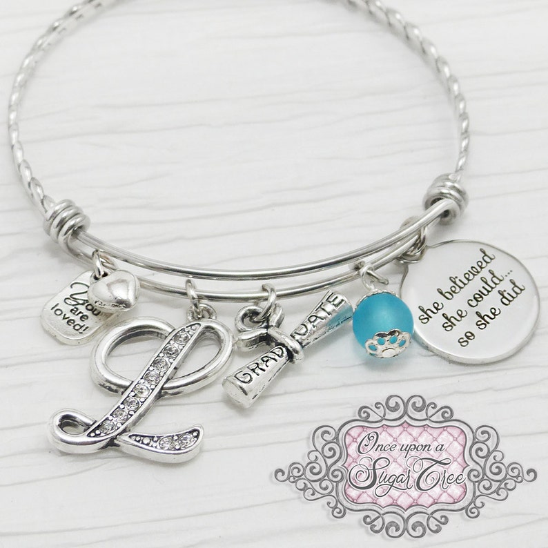 May include: Silver-toned charm bracelet with multiple pendants. Charms include a rhinestone-studded letter "L", a graduation scroll, a blue bead, and a disc with the inscription "she believed she could... so she did."
