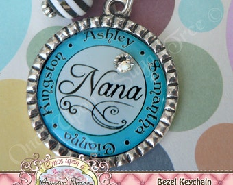 May include: A silver bezel keychain with a blue and white background. The keychain features the name "Nana" in a cursive font, along with the names "Ashley" and "Kingston" in a smaller font. The keychain also has a small rhinestone.