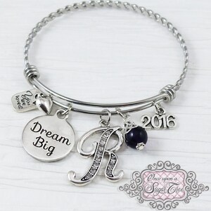 May include: Silver-toned bangle bracelet with multiple charms. Charms include a round pendant with "Dream Big" text, a letter "R" encrusted with rhinestones, a dark blue bead, a "2016" charm, and a small tag that says "You are loved!"