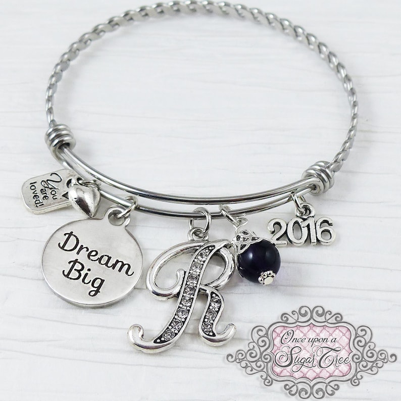 May include: Silver-toned bangle bracelet with multiple charms. Charms include a round pendant with "Dream Big" text, a letter "R" encrusted with rhinestones, a dark blue bead, a "2016" charm, and a small tag that says "You are loved!"