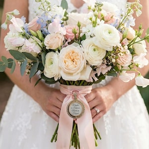 May include: A floral bouquet featuring white and pale pink roses, ranunculus, and light blue delphinium with green eucalyptus leaves. A light pink ribbon wraps the stems, accented by a circular gold charm with the text Maybe in Distance Never in Heart.