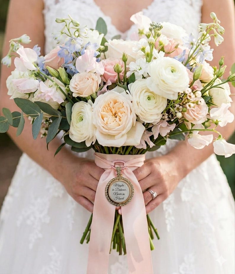 May include: A floral bouquet featuring white and pale pink roses, ranunculus, and light blue delphinium with green eucalyptus leaves. A light pink ribbon wraps the stems, accented by a circular gold charm with the text Maybe in Distance Never in Heart.