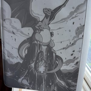 May include: A rectangular gray binder featuring a monochromatic illustration of a dragon-like creature with large wings perched atop a jagged mountain peak. The creature has a rounded belly and a long tail, with flames visible at the base of the mountain.