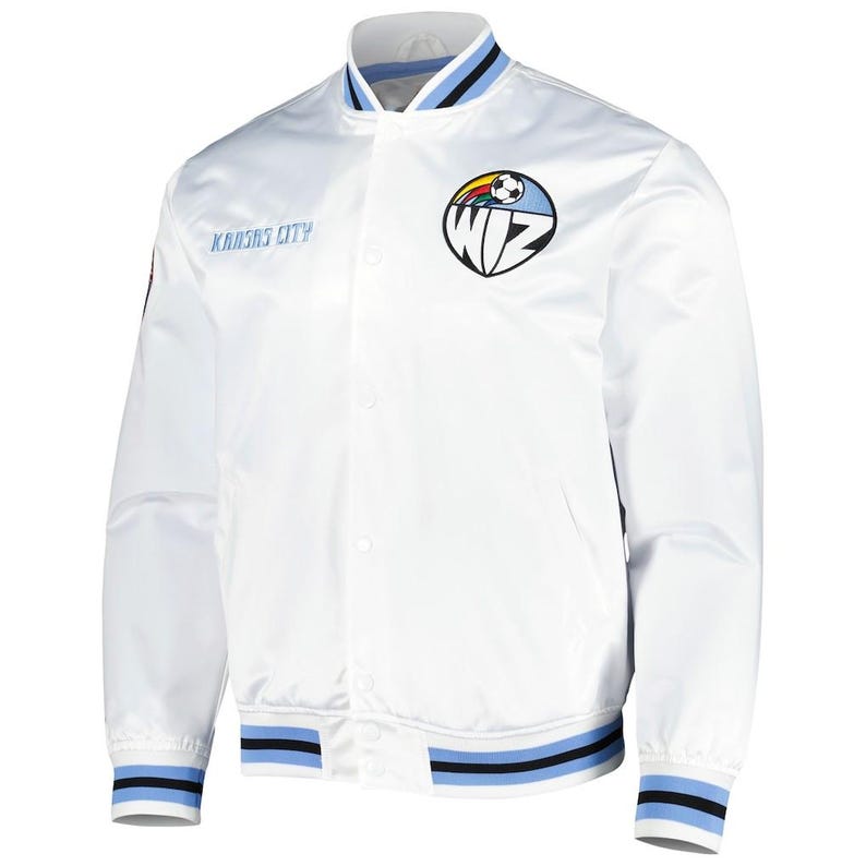 Puede incluir: Una chaqueta bomber de sat&eacute;n blanco con cierre frontal de botones a presi&oacute;n. El pecho muestra el texto KANSAS CITY en azul y un logotipo circular con la palabra WIZ, un bal&oacute;n de f&uacute;tbol y un gr&aacute;fico de arco&iacute;ris. El cuello y los pu&ntilde;os tienen rayas azules y negras.