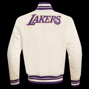 May include: Cream-colored satin jacket with purple and gold embroidered "LAKERS" lettering across the back. The jacket features purple and white striped trim on the collar, cuffs, and hem. A classic sports-themed jacket.