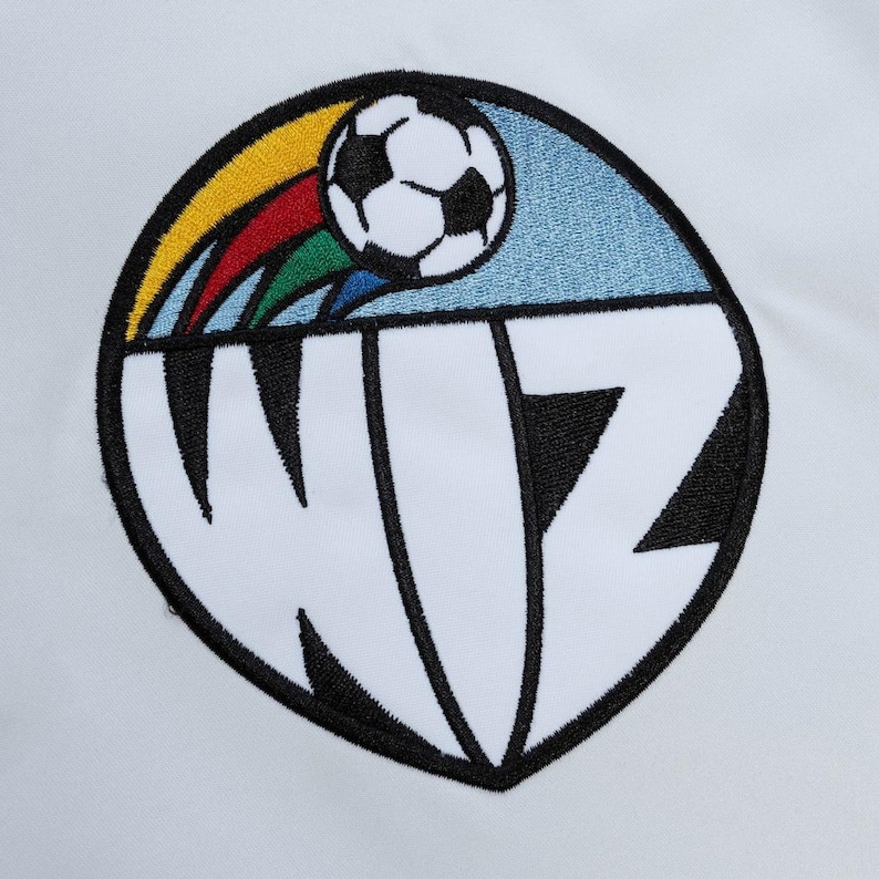 Puede incluir: Un parche bordado circular que presenta un bal&oacute;n de f&uacute;tbol en blanco y negro sobre las letras WIZ en fuente negra negrita. Un arco&iacute;ris de colores amarillo, rojo, verde y azul se extiende detr&aacute;s del bal&oacute;n sobre un fondo azul claro.