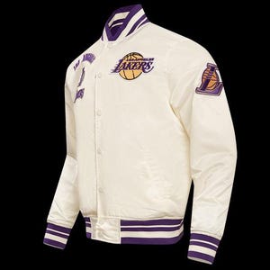 May include: Cream-colored satin jacket with purple and white striped trim. Features the Los Angeles Lakers logo and team name in purple and gold. Includes patches on the chest and sleeve with team branding. Button-up front and a classic bomber jacket style.