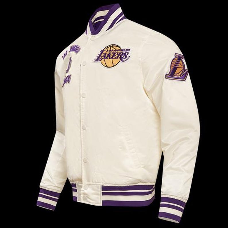 May include: Cream-colored satin jacket with purple and white striped trim. Features the Los Angeles Lakers logo and team name in purple and gold. Includes patches on the chest and sleeve with team branding. Button-up front and a classic bomber jacket style.