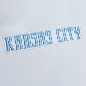 Puede incluir: Un parche de tela blanca con el texto KANSAS CITY bordado en hilo azul claro. Las letras tienen un estilo de fuente en bloque y todo el parche est&aacute; delineado con un borde blanco, creando una apariencia visual limpia y distintiva.