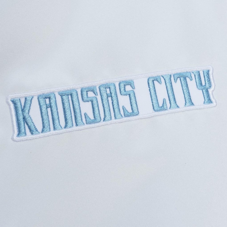 Puede incluir: Un parche de tela blanca con el texto KANSAS CITY bordado en hilo azul claro. Las letras tienen un estilo de fuente en bloque y todo el parche est&aacute; delineado con un borde blanco, creando una apariencia visual limpia y distintiva.