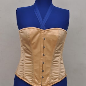 May include: A beige satin corset with a structured design, displayed on a blue mannequin. The corset features a series of metal clasps down the front and vertical seams for shaping. The garment has a curved hemline and a strapless design.