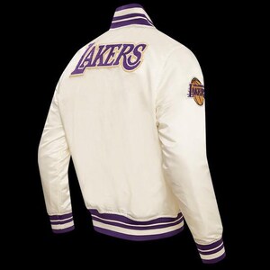 May include: Cream-colored satin jacket with purple and gold accents. The back features the word "LAKERS" in purple and gold embroidery. A basketball logo patch is on the sleeve. The jacket has a striped collar, cuffs, and hem.