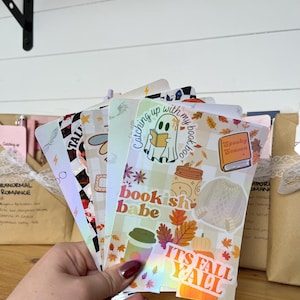 May include: A hand holds a stack of colorful, fall-themed bookmarks. The bookmarks feature various designs, including a ghost reading a book, pumpkins, and the text "bookish babe" and "It's Fall Y'all". The bookmarks have a holographic sheen.