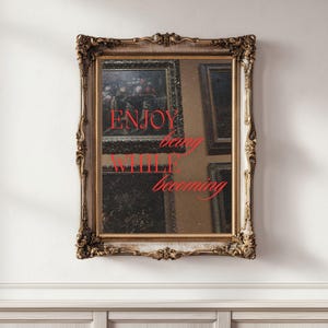 以下が含まれることがあります： 赤い文字で「ENJOY being WHILE becoming」と書かれた、装飾的な金色のフレームの鏡。鏡はアートワークを映し出し、白い壁に取り付けられています。フレームには複雑な装飾が施されています。