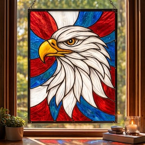 May include: Stained glass panel featuring a detailed bald eagle head with white feathers, a yellow beak, and a focused gaze. The background showcases red, white, and blue abstract shapes, reminiscent of the American flag. The panel is framed in black and hangs from a chain.