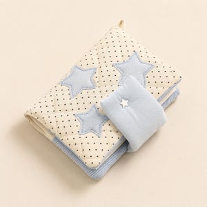 May include: A quilted fabric book cover featuring a cream-colored base with small black polka dots. The cover is decorated with three light blue fabric stars and includes a light blue flap closure secured by a small white star-shaped button.