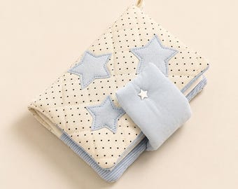 May include: A quilted fabric book cover featuring a cream-coloured base with small black polka dots. The cover is decorated with three light blue fabric stars and includes a light blue flap closure secured by a small white star-shaped button.