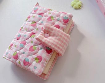 May include: A quilted fabric notebook cover featuring a pink and white gingham pattern with a strawberry print. The cover includes a matching fabric strap secured by a small pink heart-shaped button, enclosing the pages within the soft exterior.