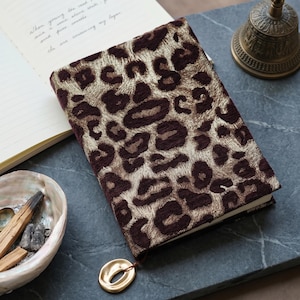 May include: A rectangular notebook featuring a textured cover with a dark brown and metallic gold leopard print pattern. The notebook includes a gold-toned oval charm attached to a thin cord at the bottom edge, providing a decorative accent.