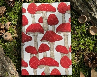 May include: A rectangular notebook with a white cover featuring a repeating pattern of red mushrooms with white spots and light brown stems. The mushrooms are arranged in a dense, vertical layout across the entire surface of the notebook cover.