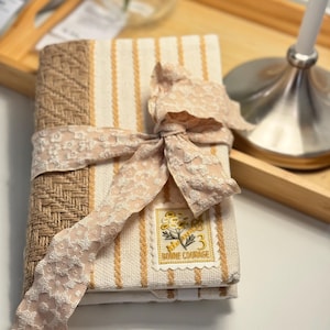 May include: A bound book with a beige and cream striped cover, tied with a light pink lace ribbon. The book has a woven brown spine. A decorative stamp with the text "Bonne Courage" is on the cover.