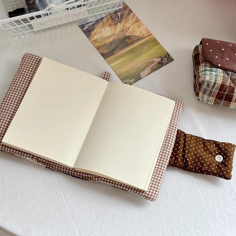 May include: An open notebook with blank cream-colored pages, encased in a brown and white gingham fabric cover. A brown fabric strap with white polka dots and a single white snap button secures the cover, which is laid flat.