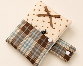 May include: A fabric pouch featuring a beige upper section with brown star patterns, a brown ribbon bow, and a small black cat embellishment. The lower section is a brown, blue, and white plaid fabric with a matching side flap secured by white snaps.
