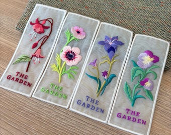 May include: Four rectangular bookmarks made of sheer fabric, each featuring colourful floral embroidery and the text THE GARDEN. The designs include a pink fuchsia, a pink poppy, a purple bellflower, and a purple pansy, all with green stems.