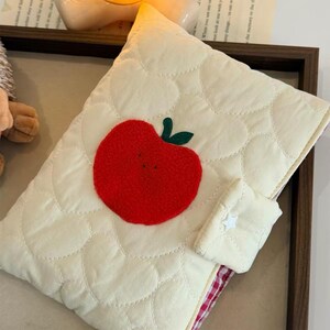 May include: A cream-colored quilted pouch featuring a repeating heart-shaped stitch pattern and a large, textured red apple appliqu&eacute; with green leaves. The pouch includes a small flap closure secured with a white star-shaped button detail.