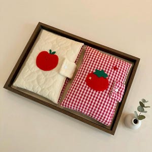 Könnte beinhalten: Zwei rechteckige Stoffhüllen für Bücher oder Tablets, eine cremefarbene mit gesteppter Textur und roter Apfel-Applikation, die andere mit rot-weißem Vichy-Muster und roter Tomaten-Applikation, beide mit kleinen Laschenverschlüssen versehen.