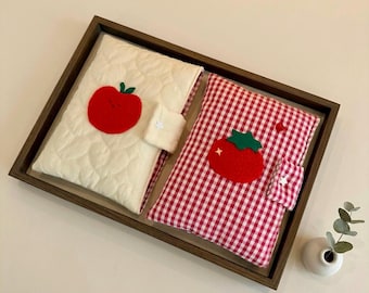 May include: Two rectangular fabric covers for books or tablets, one cream-colored with a quilted texture and a red apple appliqué, the other featuring a red and white gingham pattern with a red tomato appliqué, both with small tab closures.