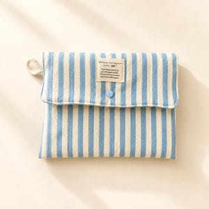Könnte beinhalten: Eine rechteckige Stofftasche mit vertikalen hellblauen und cremefarbenen Streifen. Die Klappe enthält einen kleinen hellblauen Knopf und ein rechteckiges weißes Etikett mit dem Text "Focus on your own life" und weiterem kleinem Text.